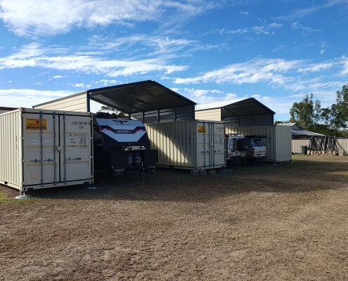 Shade sheds with parked caravan and cars inside next to shipping containers