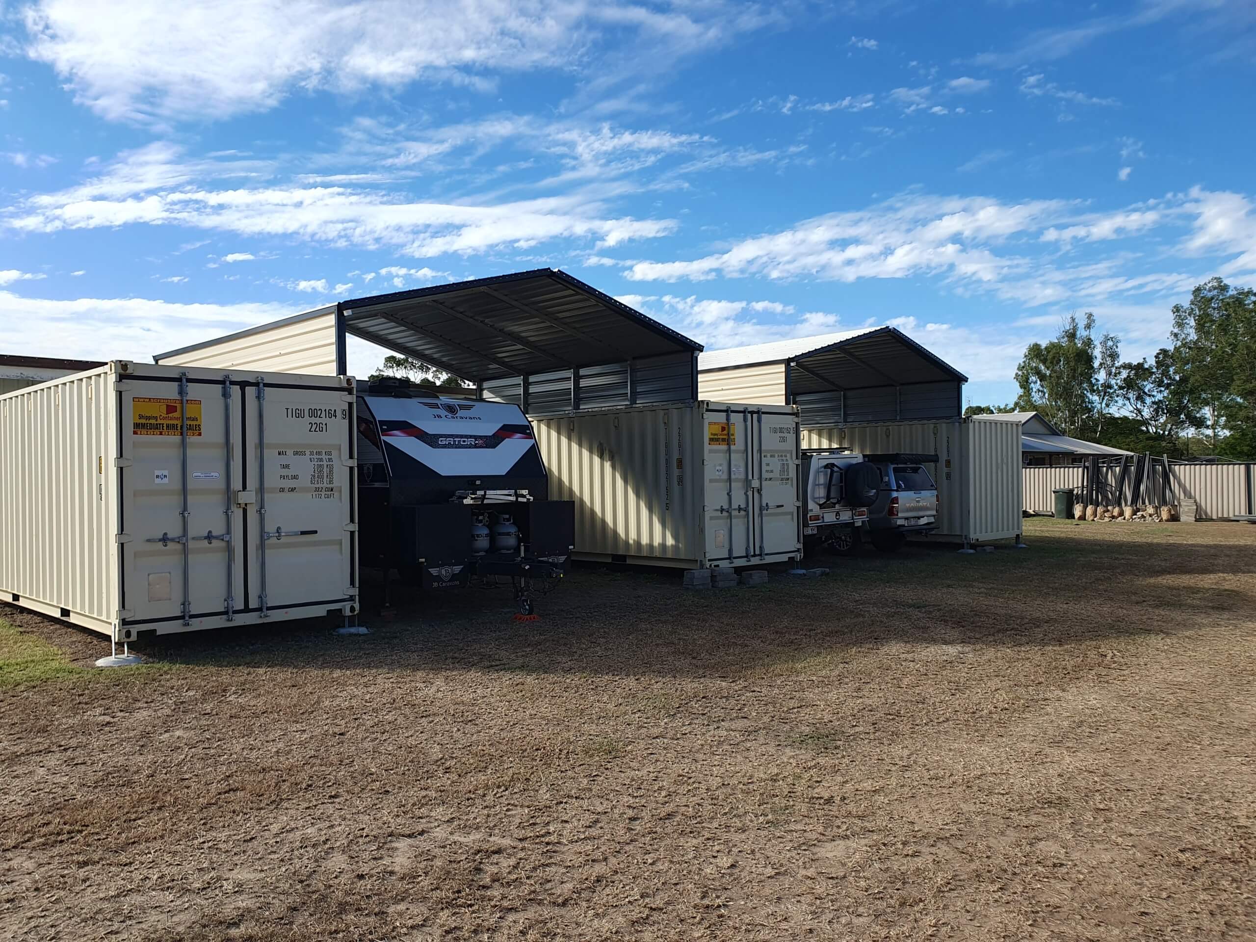 Shade sheds with parked caravan and cars inside next to shipping containers